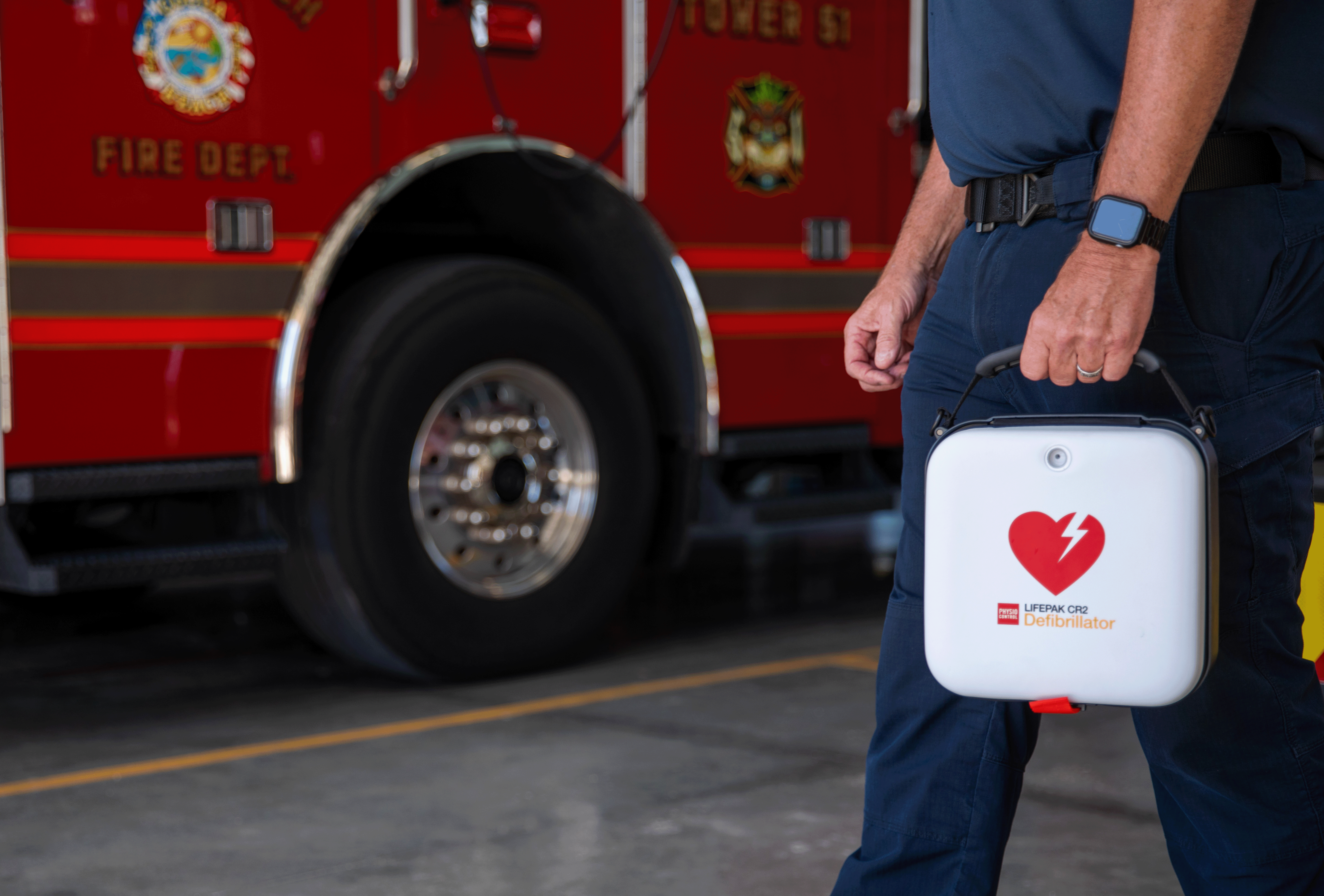 Firefighter carrying a LIFEPAK CR2 AED in front of a firetruck, demonstrating emergency preparedness and rapid response.