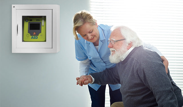 Elderly man in a retirement home receiving assistance from a nurse with an AED nearby, highlighting senior safety and emergency preparedness.